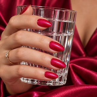 Hand mit metallic lila-roten Nägeln hält ein Wasserglas vor rotem Satin – Rockstar Nails Gellack 90012.