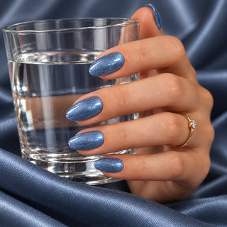 Hand mit schimmernden blaugrauen Nägeln hält ein Wasserglas vor blauem Satin – Rockstar Nails Gellack 90233.