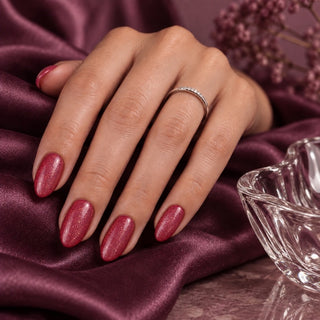 Hand mit violett-roten Glitzernägeln neben einem Glas auf violettem Satin – Rockstar Nails Gellack 90013.